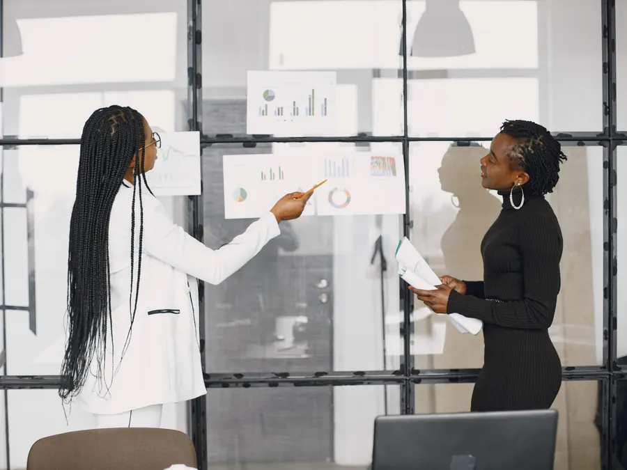 Two women working through printed charts pinned to a glass wall.