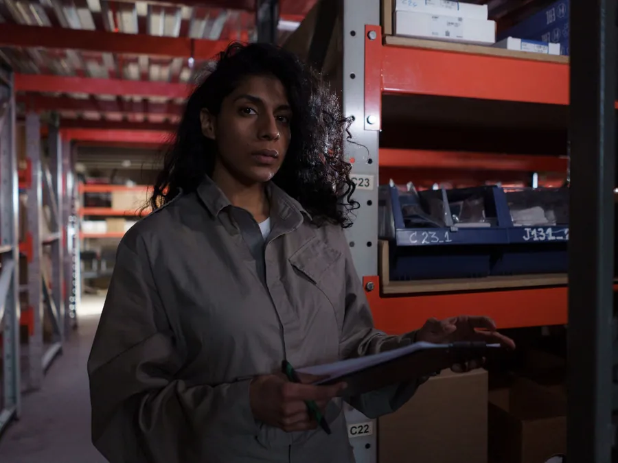 A woman with a clipboard checking stock along a warehouse aisle.